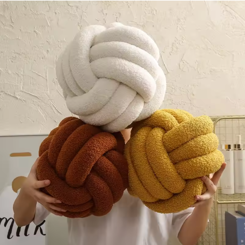 Three soft Knot Plush Pillows in white brown and yellow held by a person against a textured wall background